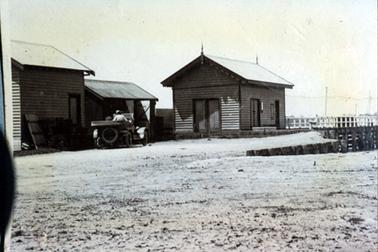 This image shows three small weatherboard sheds at the entrance to a jetty.  A small open top motor vehicle, with a driver at the wheel, is standing in front of the smaller middle shed.  A large spare tyre is attached to the back of the vehicle.  White posts with two railings can be seen on each side of the jetty.