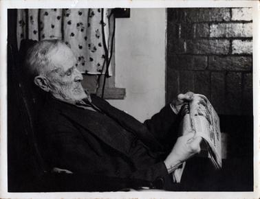 This image shows an elderly man sitting in a chair reading a newspaper.  He has grey hair and beard and is wearing a dark coat.  He is holding the paper in both hands.  There is a curtain on the wall beside him and two electrical cords can be seen on the wall.  Part of the wall on the right-hand side of the photograph is brick.  