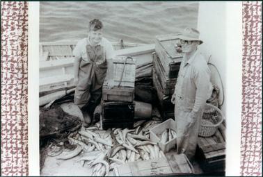 This image shows a boy and a man in a boat with a lot of fish on the bottom of the boat.  They are both smiling and looking at the camera.  The boy is wearing a short sleeved shirt, overalls and rubber boots.  The man is wearing a check shirt, cardigan and long trousers.  He is wearing a hat a dark rimmed spectacles.  There are a lot of wooden boxes in the boat, a bucket and plastic basket.  Part of a fishing net can be seen on the left-hand side of the boat.  There is an oar behind the boy.  Water is in the background.  