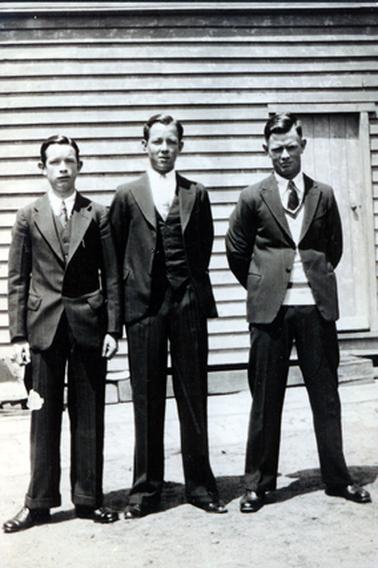This image shows three young men standing in front of a weatherboard building posing for a photograph.  They are all wearing white shirts, ties and dark coloured suits.  The two on the left are wearing waistcoats under their coats and the man on the right is wearing a light coloured pullover.  The young man on the left has his arms by his side and the other two have their arms clasped behind their backs.  None of them are smiling.  A small wooden door can be seen on the right-hand side of the building.  
