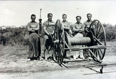 This image shows a group of five men standing behind a fire hose, arms crossed, posing for a photograph.  The hose reel has two very large wheels. There are bushes behind them and the tops of roofs can be seen in the background.  All the men have similar hair styles and none of them are smiling.  They are all wearing similar short-sleeved open-necked shirts with a logo on the front and long trousers.