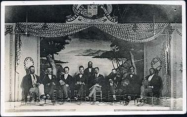 The image shows eight men seated on chairs, and one man standing, on an improvised stage. Two of the men are holding small accordions, one is holding a banjo and two are holding tambourines. The stage has a backdrop depicting an island scene, there are curtains draped above and decorative screens on either side. There is a coat of arms above the stage with the words “Hastings Minstrels Victory is in Truth”. Six wooden benches are on the floor in the foreground. The second original of this image is a Post Card.