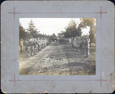 The  image shows two rows of men in uniform.  Some men on the left are playing brass instruments including trumpets and on the right some men are playing trumpets and one man is playing a large drum.  Oxen are seen standing further along the rows of men plus a man on horseback.  Pine trees are seen on both sides of the image.  The men are wearing black armbands. 