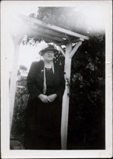 This image shows an elderly woman standing under a pergola, posing for a photo.  She is wearing a black dress, coat and hat.  She is wearing a necklace.  A brooch pinned to her left lapel.  Part of a scarf can be seen underneath her coat.  A large flowering shrub is on the right-hand side of the image and a garden can be seen behind her.  