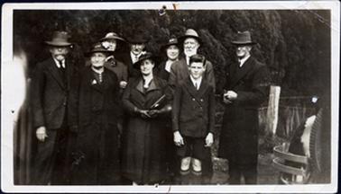 This image shows a group of four men, four woman and a boy posing for a photograph.  All the men are wearing three piece suits, ties, white shorts and hats.  The man on the far right side of the image is wearing an overcoat.  The boy is wearing a double-breasted coat, white shirt, tie, long socks and long shorts.  All the woman are well dressed with hats and coats.  There is a post and barb-wire fence behind them and thick trees behind the fence.  