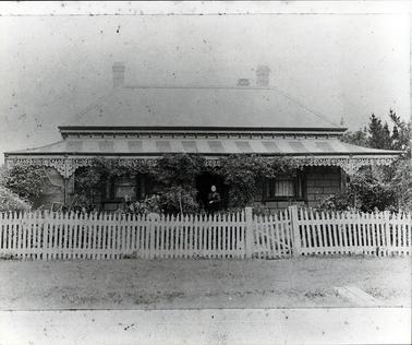 This image shows a cottage with a lady standing in the doorway.  It is of brick construction with a galvanized iron roof.  A lower roof is covering a verandah which is across the entire width of the cottage.  There is a wrought iron lace strip across the verandah and on the verandah posts.  Creepers are growing up the verandah posts.  Three windows can be seen at the front of the cottage and three chimneys can be seen on the roof.  A picket fence with a little gate is across the front of the cottage with a garden behind it.  