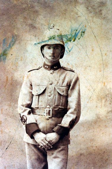 This image shows a young man in full military uniform, standing posing for a photo.  He is wearing a helmet with a chin strap and his jacket has large buttons, two large pockets and a wide belt.  His collar and cuffs are a dark colour.  His hands are clasped at the front.  