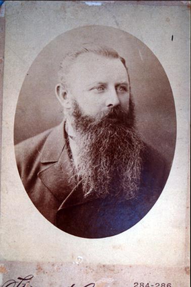 This image shows an oval portrait of a man, taken from the chest up.  He has a greying beard and moustache and his hair is neatly combed back.  He is looking away from the camera.  He is wearing a double breasted coat.  Part of a white shirt with a small high collar can be seen beneath the beard.  
