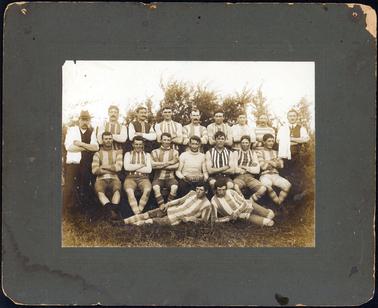 This image shows a football team posing for a photograph.  There are nine men standing at the back, seven men sitting on chairs in the middle and two men half lying on the grass at the front.  The two men standing at either end of the back row are not dressed in football clothes.  They have white shirts, rolled up to the elbow, vests and have a fringed cloth over one shoulder. The man sitting in the centre is holding a football on which is written: “Premiers 1910”.  Small trees can be seen behind the men.  