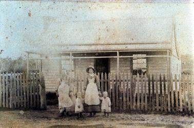 This image shows a weatherboard house with a four posted verandah across the front.  An open door is in the centre of the building with a set of windows on either side.  A picket fence is across the front with a small open gateway.  A man, woman and two small children are standing in front of the fence, posing for the photo.   