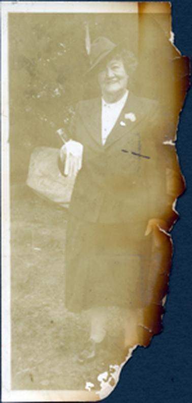 This image is a full length photo of a woman posing for a photograph.  Half of the photograph has been burnt and torn away.  The woman is wearing a two piece suit with a high necked white blouse.  She is wearing a hat, tilted to the side and has a smile on her face.  A brooch in the shape of Australia is on her left lapel and white gloves can be seen in her right hand.  A large rock appears to be behind her and part of a tree can be seen behind that. 