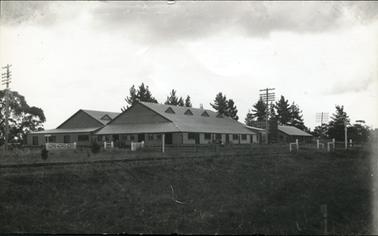 This image shows two large sheds with another smaller building on the right-hand side of the photograph.  There is a tall box-like structure between the large shed and the smaller building.  A vehicle can be seen parked beside the large shed. Three tall electricity poles can be seen.  A railway line runs in front of the buildings. A post and rail fence can be seen along with white posts, gates and signs for the railway.  A grass paddock is in the foreground and tall trees can be seen behind the buildings.  There are clouds high in the sky.