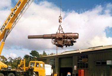 This image shows a gun barrel being lifted in the air by a crane. The crane has ‘Tolson’s’ written along the side and ‘Tolson’s Mobile Cranes’ is printed on the truck.  A large galvanised iron shed is on the right-hand side of the image.  A man can be seen bending down in front of the partly opened door.  