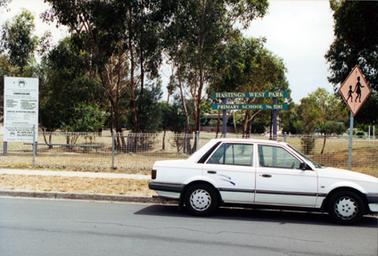 This image shows a school grounds with the sign “Hastings West Park Primary School No. 5202” behind the front fence.  A white notice board showing the school’s curriculum is on the far left-side and a children’s road safety sign is on the far right-hand side of the image.  Tall trees are scattered around the grounds.  A wire fence runs across the front of the grounds and a concrete footpath and a street are in the foreground.  A white motor car is parked on the street.  