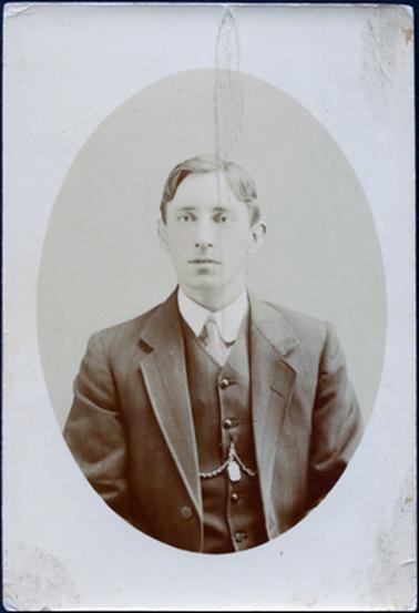 This is an oval image, from the waist up, of a young man dressed in a three piece suit with a tie.  The collar on his shirt is rounded.  He has a chain on his waistcoat which is buttoned up.  His hair is straight and neatly parted on the left-hand side.  
