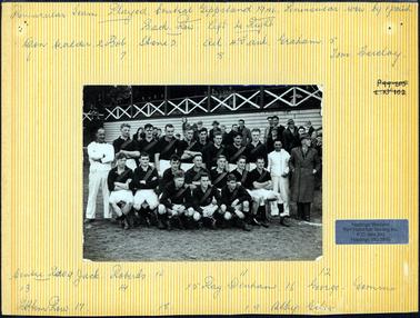 This image shows a football team with their supporters and spectators.  The footballers are in three rows, eight in the back row, seven sitting in the middle row and three crouched in the front row.  They have long-sleeved dark shirts, white shorts and a sash across their shoulder to the waist.  Two men, wearing all white are standing on either side of the team and a man wearing an overcoat is standing on the far right-hand side of the photograph.  There are spectators behind the players and more can be seen in the stand behind them.  The stand is wooden with a wooden railing with cross pieces of timber in each section.  There is grass in the foreground.  