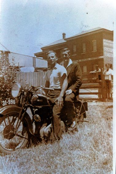 This image shows two men sitting on a motorcycle posing for a photo.  The man on the front of the bike is wearing a white short-sleeved shirt and dark trousers.  The man behind him is wearing a suit with an open-necked white shirt.  The bike has the registration number plate VIC 31-996 above the front wheel.  Two people are leaning on a fence behind the bike looking on.  A two story building with ‘Royal Hotel’ on the front is in the background.  ‘Fosters Lager’ is painted on the side of the hotel.  Part of a wooden fence and an awning can be seen on the left-hand side of the image.  
