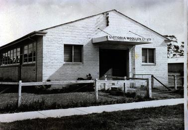 The image shows part of a large brick building with ‘school type’ windows along the left-hand side of the photograph and two sets of louvres either side of the wide front door.  On the awning over the doorway is the sign VICTORIA WOOLLEN Co Ltd HEAD OFFICE THOMAS ST YARRAVILLE. There is a post and wire-netting fence across the front of the building.  A concrete footpath and grass are in the foreground.  Part of another building can be seen in the distance on the right-hand side of the photograph with a large tree behind it.  