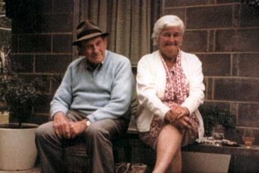 This image shows an elderly man and woman sitting on a bench in front of a brick wall.  They are both smiling for the camera.  He is wearing a grey pullover, brown trousers and brown hat.  The woman is wearing a white cardigan over a pink and white print dress.  She has white hair.  A carry bag is on the ground between them.  Two half filled glasses are sitting on the bench beside the woman.  A window with a brown curtain is behind the couple.  Two large pots with plants are on either side of the couple. The photograph has been laminated onto a white A4 page.  Printed in black ink beside the photograph is: “Noon & Bill Patience Skinner St Hastings Melbourne Cup Day 1980”.
