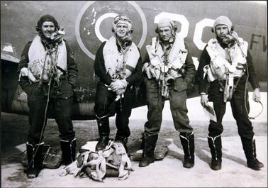 This image shows four airforce men standing in front of a plane posing for a photograph.  They are dressed in full operational gear.  The man second from the left has his foot on top of some gear on the ground.  Only part of the plane can be seen behind them.