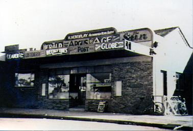 This image shows a brick shop front with an awning over the footpath.  Large windows are on either side of the front door and a set of small windows are above.  The signage at the top of the building is in three layers.  The back section reads: “M. Mentiplay Aurthorized NewsAgent”.  The middle section reads: “Herald Read The Argus Age The Sun”.  The front section reads: “Leader Listeners Weekly Times Post Globe”.  An advertising board is leaning against the shop on the footpath and two bicycles can be seen at the right-hand side of the footpath.  
