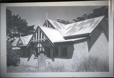 This image shows the front of a building with a corrugated iron roof. Three triangular pediments can be seen on the roof line as well as three chimneys.  Three sets of windows are either side of the wooden front door and three small gable finials on the roof can also be seen.  Untidy grass is at the front of the building and tall pine trees are in the background.  Part of a bushy tree is on the far left-hand side of the image.  Written on at the bottom of the image is: “State School No 841, Flinders 1970”.