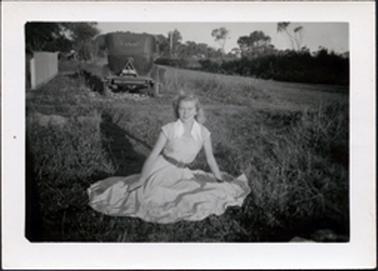This image shows a young woman sitting on the grass posing for a photo.  She is wearing a light coloured dress with a flared skirt spread out in a circle on the grass. The dress has a double white collar and she is wearing a dark coloured belt.  Her blonde hair is back behind her ears.  An old 28 model car with the number plate 83.381 is parked on the footpath behind her.  A street, which is lined with trees and shrubs is on the right-hand side of the image and part of a picket fence can be seen on the left-hand side of the image.  