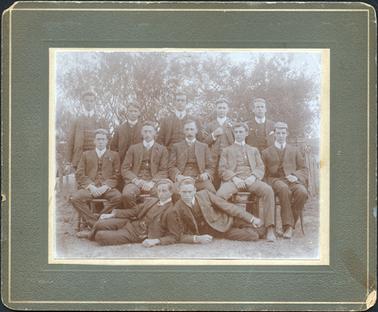 This image is of a group of men posing for a photograph.  They are in three rows.  Five are standing at the back, five are seated in the middle and two are ‘lounging’ on the ground at the front.  They are all dressed in three-piece suits and have fob chains.  They are wearing very small caps on their heads.  There are trees in the background as well as an old fence.  