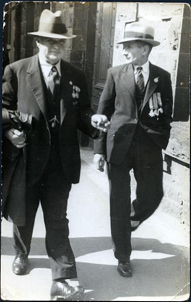 This image shows two men walking along a footpath.  They are both dressed in three piece suits, white shirts, ties and hats.  The man on the left-hand side is carrying a coat over his arm.  They are both wearing  war service medals and RSL badges.  The man on the right-hand side is looking sideways.  A small brick retaining wall and buildings can be seen behind them. 