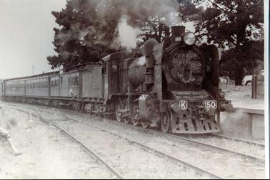 This image shows a stream strain stopped at a station.  There are three passenger carriages and part of the guard’s van can be seen at the rear.  A boy is hanging on to the outside of the first carriage.  Part of another boy is stepping into the picture on the far left-hand side.  Steam is escaping from the engine.  The letter ‘K’ and the number ‘150’ is on the front of the engine.  Printed across the front of the engine is: ‘Cannon Ball 6 R.I.P.’.  A second rail track is in the foreground and tall trees are behind the train.  Part of the platform, a fence and a motor vehicle can be seen on the far right-hand side of the image.  