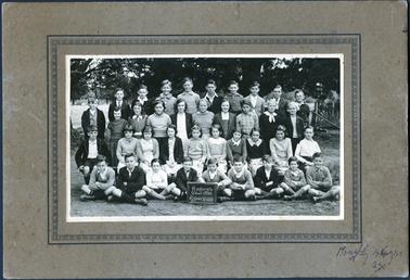 This image shows a group of school children, in four rows, posing for a photograph.  There are ten children standing in the back row, nine children standing in the second back row, ten children sitting on stools in the second front row and nine children sitting cross-legged on the ground in the front row.  A boy in the front row is holding a small blackboard which reads: “Hastings School 1098 Grades V to VIII 1937.  There are tree branches in the background.  Written in biro on the mounting is: “Roughly 44 yrs ago”.