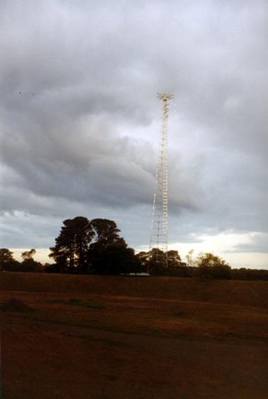 This photograph shows a tall communications tower in the centre of the image.  The roof of a building can be seen to the left of the tower.  Tall trees are on either side of the tower.  Thick grey clouds are in the sky and uneven brown coloured earth is in the foreground.  
