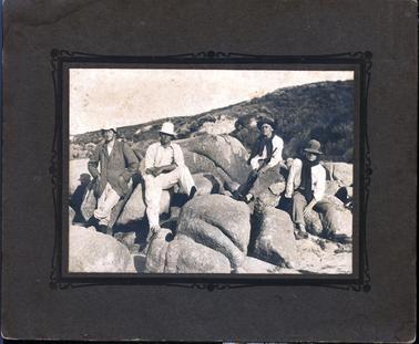 This image shows four young men sitting on large rocks with sand dunes in the background.  The man on the left-hand side has his eyes closed and his hands are in the pockets of his coat.  The man next to him is dressed all in white and has one foot up on a rock.  The other two men are dressed in white shirts, darker trousers and have a dark coloured jumper tied around their necks.  The man second from the right is holding a rifle.  All men are wearing hats.  