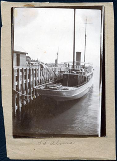 This image shows a boat tied up to a wharf.  The boat has two high masts and a large funnel.  There is a large flag flying on a flag post at the front of the boat.  The wharf is of wooden construction and part of a shed with an open door can be seen on the left of the photograph.  Trees can be seen in the far background.  At the bottom of the mount is written S S Alvina.