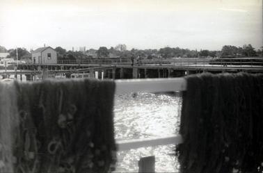 This image shows a pier with three people standing together in the centre of the image.  A shed is on the left-hand end of the pier.  Fishing nets are handing on a rail in the foreground.  A small section of the upper and lower rail can be seen between the nets.  A part of several boats is visible above the nets and motor vehicles are parked in front of the shed on the far left-hand side.  Buildings and trees are in the background.  