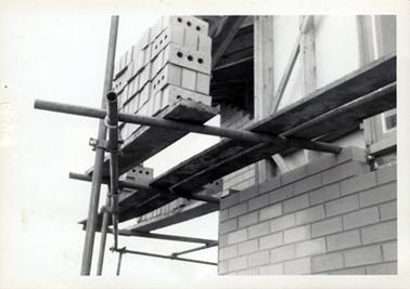 This image shows a construction site.  Scaffolding is attached to a brick building.  A pile of bricks are sitting on a plank on the scaffolding ready to be laid.  