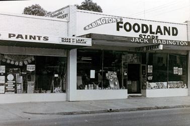 This image shows a shop frontage.  The store has a glass window front with a door in the centre.  There is a large display in the window with several small advertising posters attached to the window.  The sign “Self Service” is above the doorway.  The top section of the shop has large signage which reads: “Babington’s Foodland Store Jack Babington”.  A paint shop is attached to the left-hand side of the store.  A concrete footpath and bitumen street is in the foreground.  