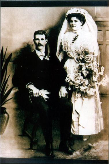 This image shows a bride and groom posing for a photo.  The groom is sitting in a wooden carved arm chair.  He is dressed in a dark three-piece suit.  His white shirt has a high collar with a small bow tie.  Both arms are resting on the arms of the chair.  He is holding white gloves in his right hand and has an orange blossom sprig on his left lapel.  His hair is parted on the left and he has a moustache.  The bride is wearing an elaborate ankle length white dress with a lace bodice, high lace collar and long tight sleeves.   She has a lace panel down the front of her dress.  She is holding a very large bouquet of flowers in her left hand. She has a headpiece and a long veil.  Her right hand is resting on the back of the chair.  A tall pot plant is to the left and a rug is on the floor to the right.  Part of a panelled wall can be seen behind the bride.  