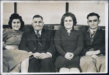 This image shows a group of four people sitting on a sofa, posing for a photo.  They are all smiling for the camera.  The young woman on the left is wearing a long sleeved dress with pin tucks above and below the waist line.  She is wearing a necklace and has thick, black wavy hair.  The man beside her is older and is wearing a three piece suit, white shirt, tie and a fob chain.  The older woman beside him is wearing a dark coloured, long sleeved dress with a brooch at the neck.  She has dark, thick hair and her hands are clasped on her lap.  The young man on the right is wearing a dark jacket, light trousers, shirt and tie.  He has black wavy hair.  Part of the ‘dots and lines’ pattern on the sofa can be seen behind them.  