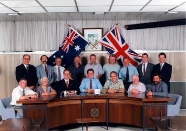 This image shows a group of fifteen men and one woman sitting and standing behind a desk posing for a photo.  The curved desk is polished timber and has a round plaque on the front.  Everyone is smiling for the camera.  Nine of the men are standing.  Everyone is well dressed.  Some men are wearing suits.  Two flags are crossed behind the group in front of a photo of the Queen.  Curtains are across a set of windows in the background above a timber panelled wall.