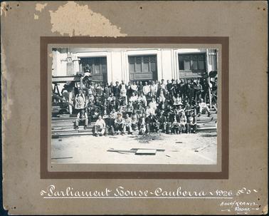 This image shows a group of approximately seventy men and one woman standing and sitting on the steps of a building.  The one woman is standing in the back row on the left-hand side of the group.  She is wearing a black hat and an overcoat with a black collar.  Most of the men are either wearing or holding a hat or cap.  Some men are dressed in three-piece suits and ties, some in working clothes and some with aprons. There are steps right across the front of the building and three large sets of windows are behind the men.  Pieces of timber are strewn across the ground in the foreground.  “Parliament House ~ Canberra ~ 1926.” is printed in white on the mounting board below the photograph.  The photographers details are also printed on the mounting board.  