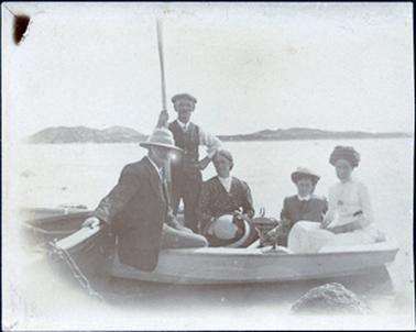 This image shows two men and three women in a boat posing for a photograph.  The man on the left is wearing a coat and hat and has his right hand resting on the side of another boat.  This boat appears to be chained to a rock which is the foreground.  The other man is standing.  He is holding an oar in his right hand and his left hand is on his hip.  He is wearing a cap, long sleeved white shirt, tie and vest with a fob chain across his chest.  The three women are very well dressed.  The woman in the centre of the boat is holding a large hat on her lap and the other two women are wearing small hats.  Land can be seen in the background.  