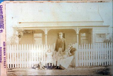 This is a very faded image of a small wooden cottage with an iron roof.  A man, woman, two small children,  a baby and a dog are outside the white picket fence on the footpath, in front of the cottage.  The woman is sitting on a chair nursing the baby.  A three post verandah in at the front of the cottage and wrought iron lace work is between the posts.  A window can be seen either side of the front door and a smaller window, with curtains, is on the right-hand side of the cottage.   The original photographers details are shown at the left-hand side.  