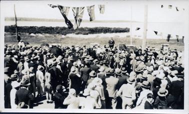 This image is of a very large gathering of men, woman and children with their backs to the camera.  A man in a three-piece suit can be seen, in an elevated position, in front of them.  All the people appear to be very well dressed and all are wearing hats.  Some umbrella’s can be seen.  A group of boys in uniform are standing in a line on the right-hand side of the image.  Two are holding flags.  There is a tall, thick post on the right-hand side with a thinner post behind it.  There are two lines of bunting attached to the posts and a line of flags is in the top centre of the image.  The foreshore is seen in the background.