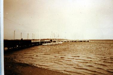 This image shows a jetty jutting out into a bay with rippling waves coming onto the foreshore in the foreground.  Three boats with tall masts are tied up on the right-hand side of the jetty and five tall masts can be seen on the left-hand side.  Land can be seen on the left-hand side in the distance.