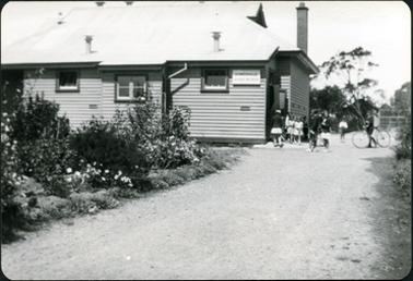 This image shows a wooden building with a sign on the wall which reads “Somerville School No. 2656”.  The building has an galvanized iron roof and a large chimney can be seen on the right-hand side of the building.  Three windows are along the wall and a group of children are standing around an open doorway on the right-hand side of the building.  One boy is standing beside a bicycle.  A large flower garden is beside the road leading into the school and trees can be seen in the background.  