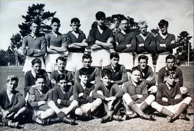 This image shows a young football team posing for a photograph.  They are all dressed in a uniform of dark coloured long sleeved shirts and white shorts. There are seven players standing in the back row, six squatting in the middle row and seven sitting cross-legged on the grass in the front row.  They all have their arms crossed.  There are large trees behind them in the background and a white goal post can be seen on the far right-hand side.  