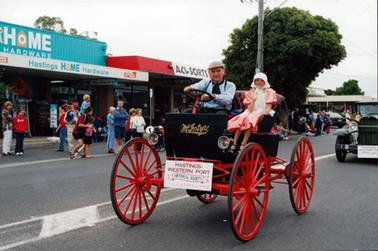 This image shows a vintage motor vehicle driving down the middle of the street.  The man driving the car is dressed in vintage clothes.  He is wearing a blue, long-sleeved shirt, wide tie, braces and a cap.  A little girl is sitting beside him, also wearing vintage clothes.  She has a pink dress with big puff sleeves and a white bonnet with a long tie at the neck.  She is waving and smiling for the camera.  The car is black with red wheels.  Written on the front in gold lettering is: “McIntyre”.  A sign is tied to the front of the vehicle which reads: “Hastings-Western Port Historical Society Incorporated”.  Part of another vintage motor vehicle with a similar sign at the front is following.  People and shops line the street on the left-hand side.  The shop that the vehicle is passing is “Hastings Home Hardware”. 