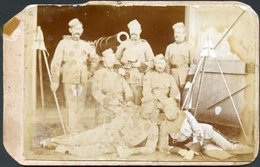 This image shows seven soldiers posing for a photograph in the doorway of a shed.  Three men are standing, two sitting on chairs and two half-lying on the ground at the front.  There is a large gun barrel behind the men.  Three small flags on poles are leaning together on either side of the men.  All men are in military uniform which includes pill-box hats, wide leather belts and a wide stripe down the side of the trousers.  A large wooden door is on the right-hand side of the photograph.  