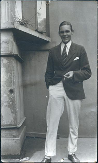 This image is a portrait of a young man standing.  He wearing a dark blazer with a handkerchief in the pocket, white shirt and striped tie and light coloured trousers.  His right hand is in his trouser pocket and his left hand is holding a cigarette.  He has a smile on his face and his hair is short and neatly parted on the left-hand side. Part of a building on the left-hand side of the photograph.  